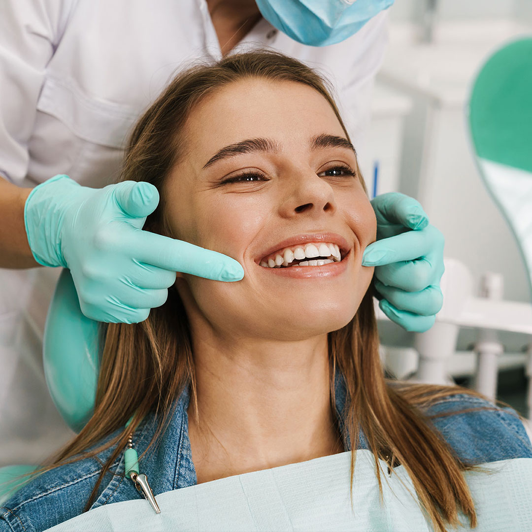 A woman receiving dental care, smiling broadly with her eyes closed, while seated in a dental chair with a dental hygienist behind her.