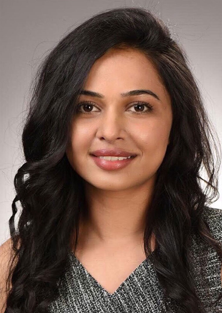 The image shows a woman with dark hair smiling at the camera. She has fair skin and is wearing a sleeveless top. Her background is plain, focusing attention on her.