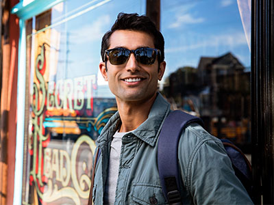 A man with sunglasses stands confidently outside a storefront, wearing a backpack and jacket, against a backdrop of a window display.