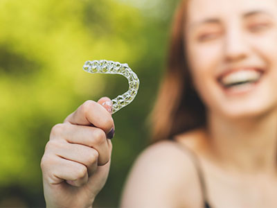 The image shows a smiling person holding a clear plastic mouthguard with their right hand against a blurred background.