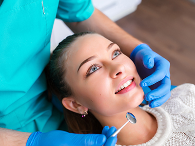 In the image, a woman is seated with her eyes closed while receiving dental care from a professional wearing blue gloves and a white mask.